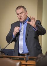 Representative Baker stands at his desk in the House Chamber and speaks into a microphone to someone out of view while gesturing with one of his hands.